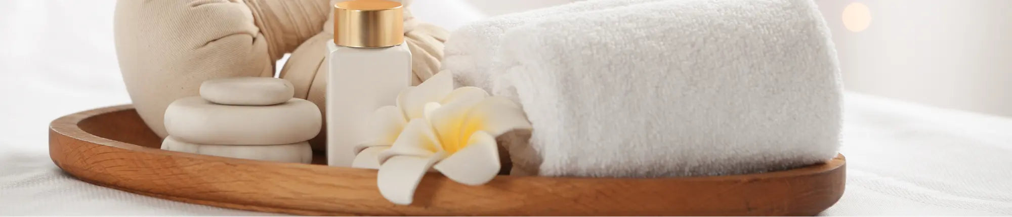 A wooden tray with stacked massage stones, a small bottle with a gold lid, a rolled white towel, and a white frangipani flower, all arranged for a spa setting.