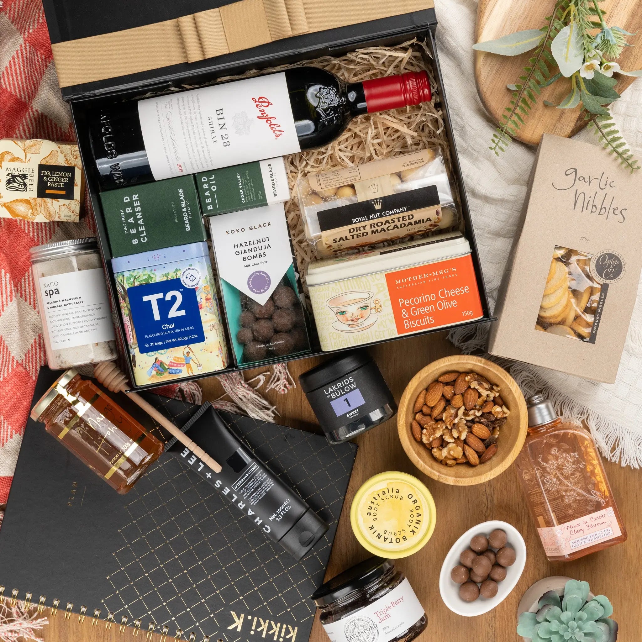 A gourmet gift box with red wine, chocolates, teas, crackers, nuts, honey, garlic nibbles, skincare products, and a notebook, arranged on a table with a red checked cloth and eucalyptus leaves.