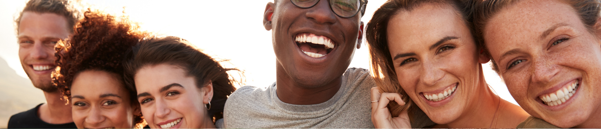 A group of six young adults stand close together outdoors, smiling and laughing at the camera in bright sunlight.