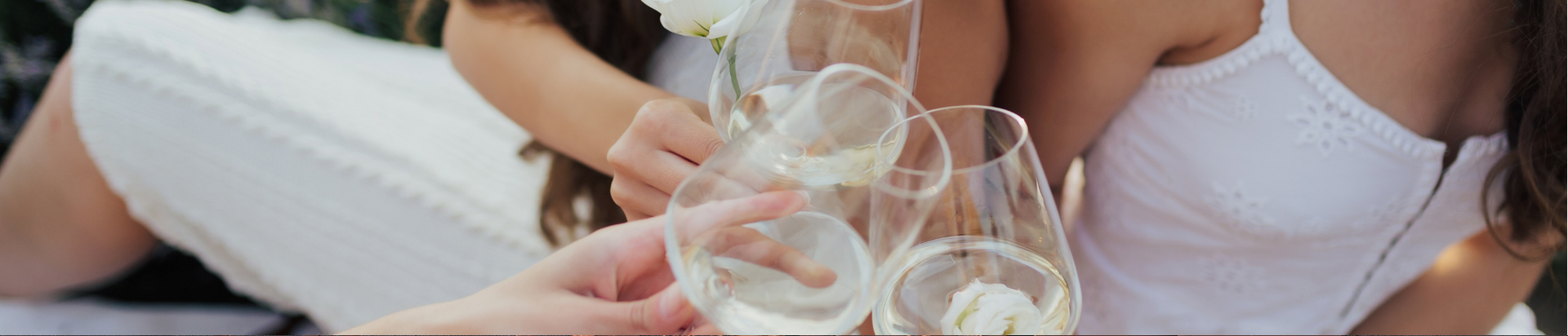Three women in white dresses clink wine glasses together, celebrating outdoors. The focus is on their hands, glasses, and white dresses, with a soft, summery atmosphere.