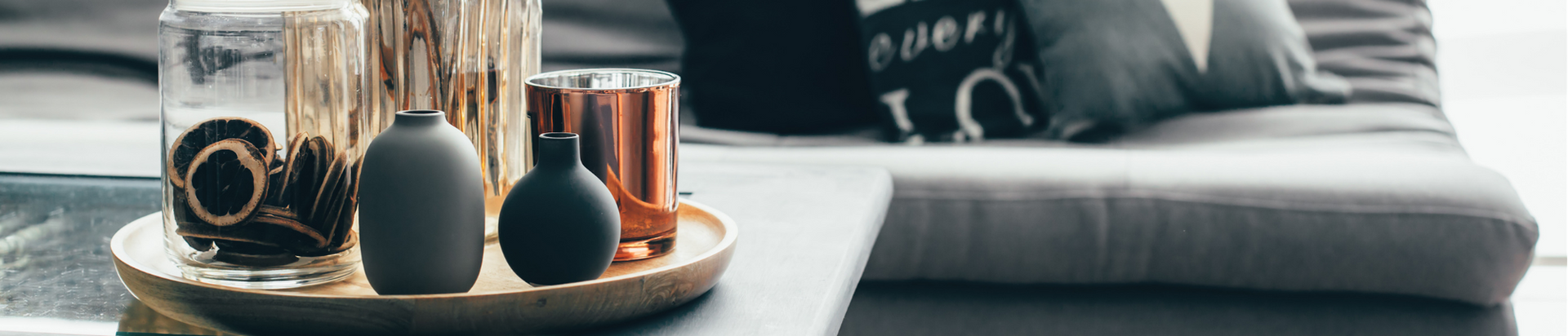 A close-up of a wooden tray on a coffee table holding two small vases, a glass jar with dried slices, and a metallic candle holder, with a grey sofa and cushions in the background.