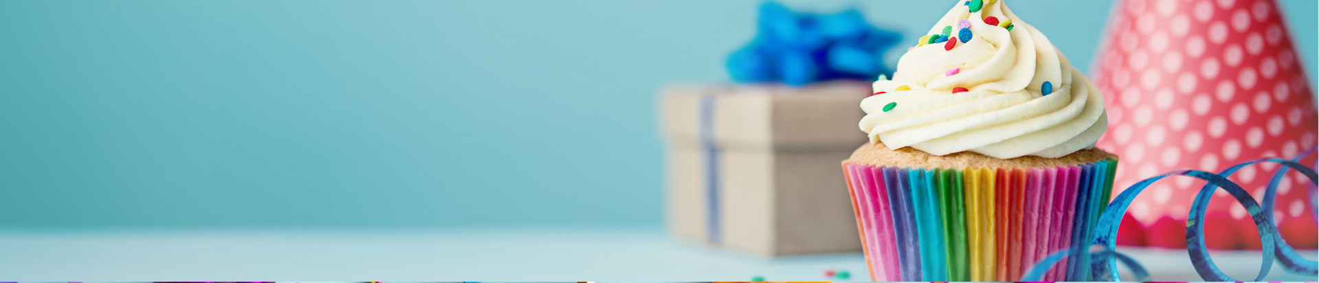 A cupcake with white frosting and sprinkles in a rainbow wrapper sits on a table, with a gift box, blue ribbon, and a pink polka dot party hat in the background.
