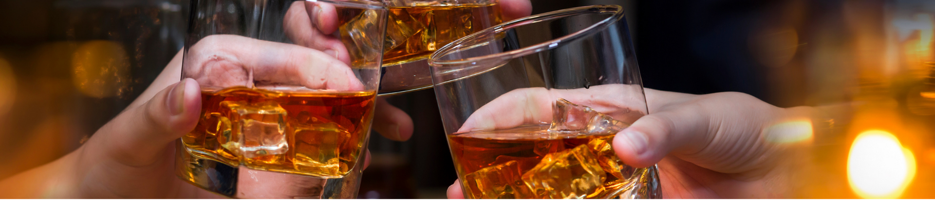 Three hands clinking glasses filled with amber whiskey and ice cubes, celebrating together against a warmly lit background.