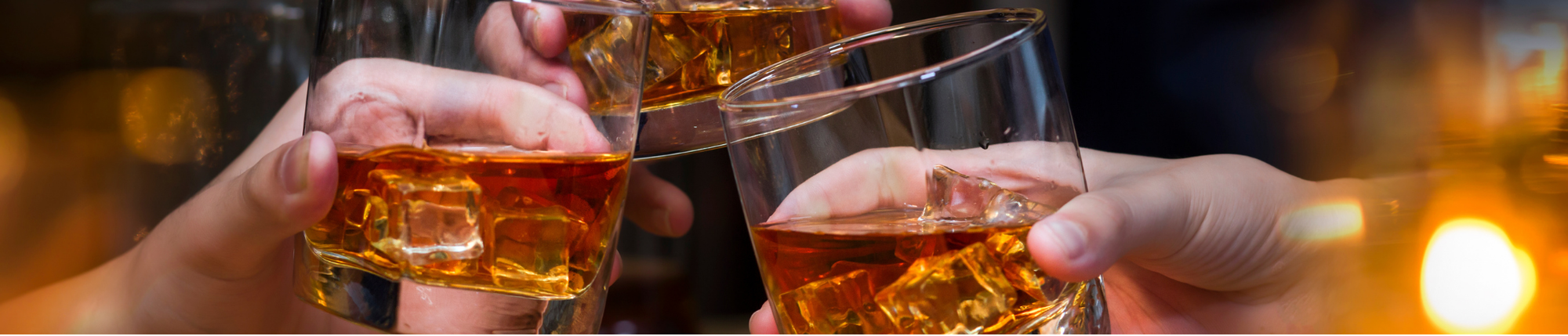 Three hands clinking glasses filled with amber whiskey and ice cubes, celebrating together against a warmly lit background.