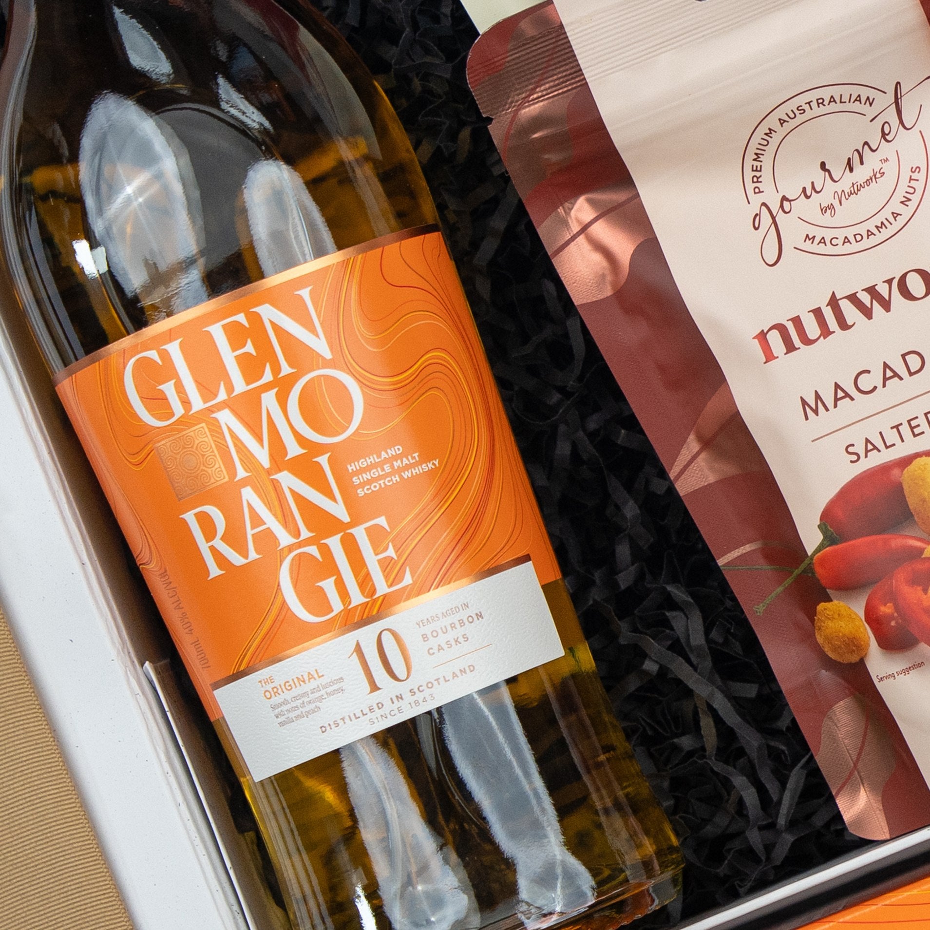 The Hamper Boutique Co Glenmorangie Moments Hamper features a Glenmorangie 10-year-old whisky in a gift box paired with Nutworks salted macadamia nuts on black shredded paper.