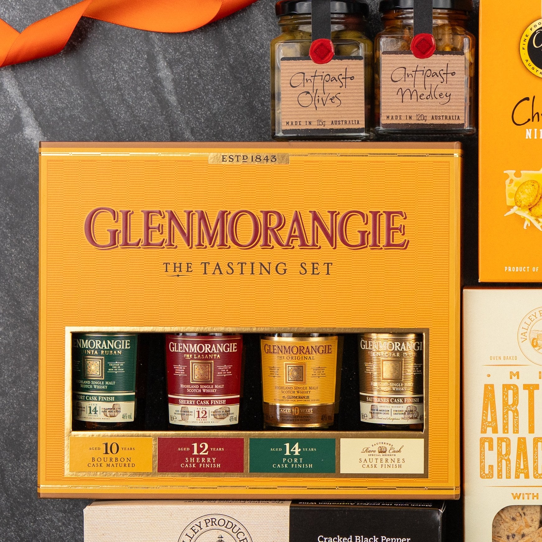 The Hamper Boutique Co Glenmorangie Tasting Experience Hamper features four whisky miniatures displayed on a dark surface with jars of antipasto, crackers, and other gourmet treats.