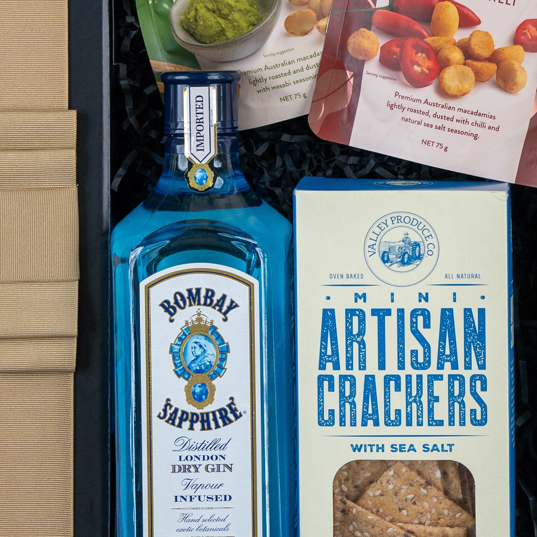 The Hamper Boutique Co’s Bombay Sapphire Botanical Escape Hamper features a blue bottle of Bombay Sapphire gin, artisan crackers, and snack packs with macadamias and tomatoes in a gift box finished with a gold-striped ribbon.
