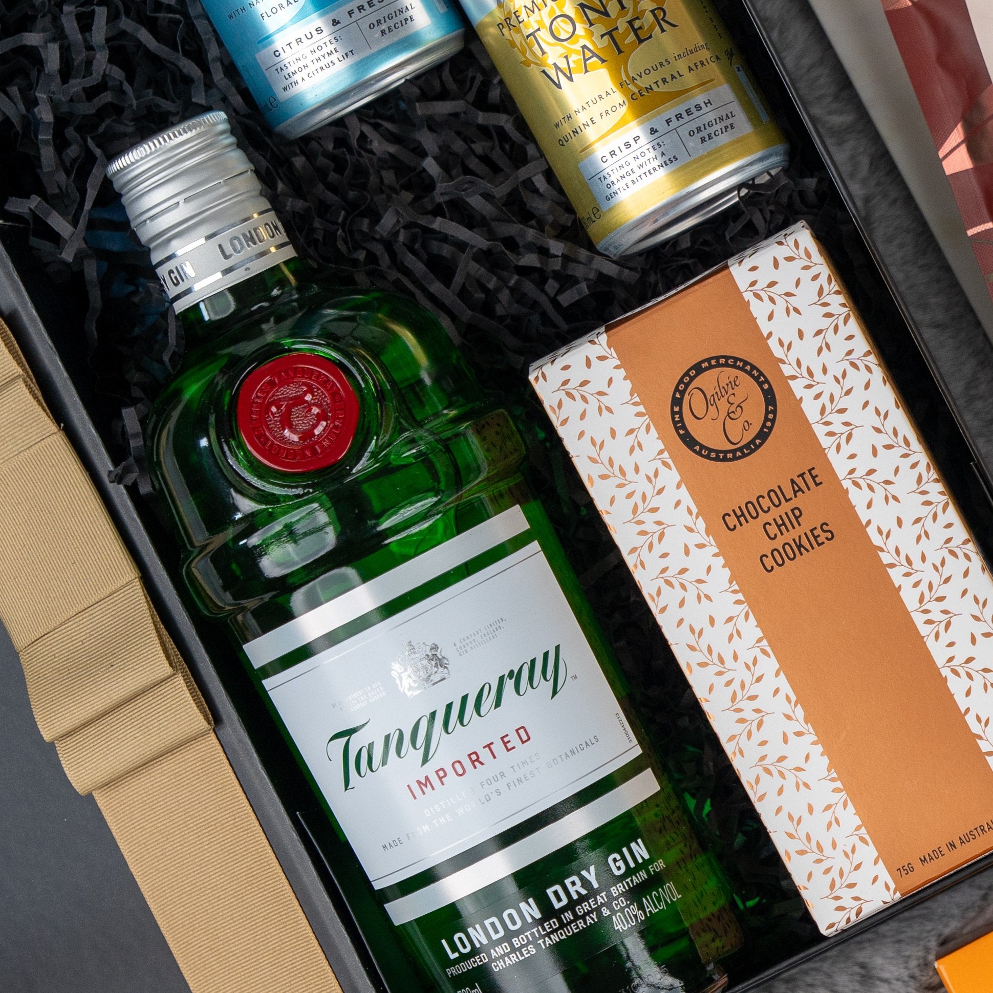 The Hamper Boutique Co Tanqueray Gin Journey Hamper includes Tanqueray London Dry Gin, chocolate chip cookies, and two artisan tonic water cans, beautifully arranged on black shredded paper.