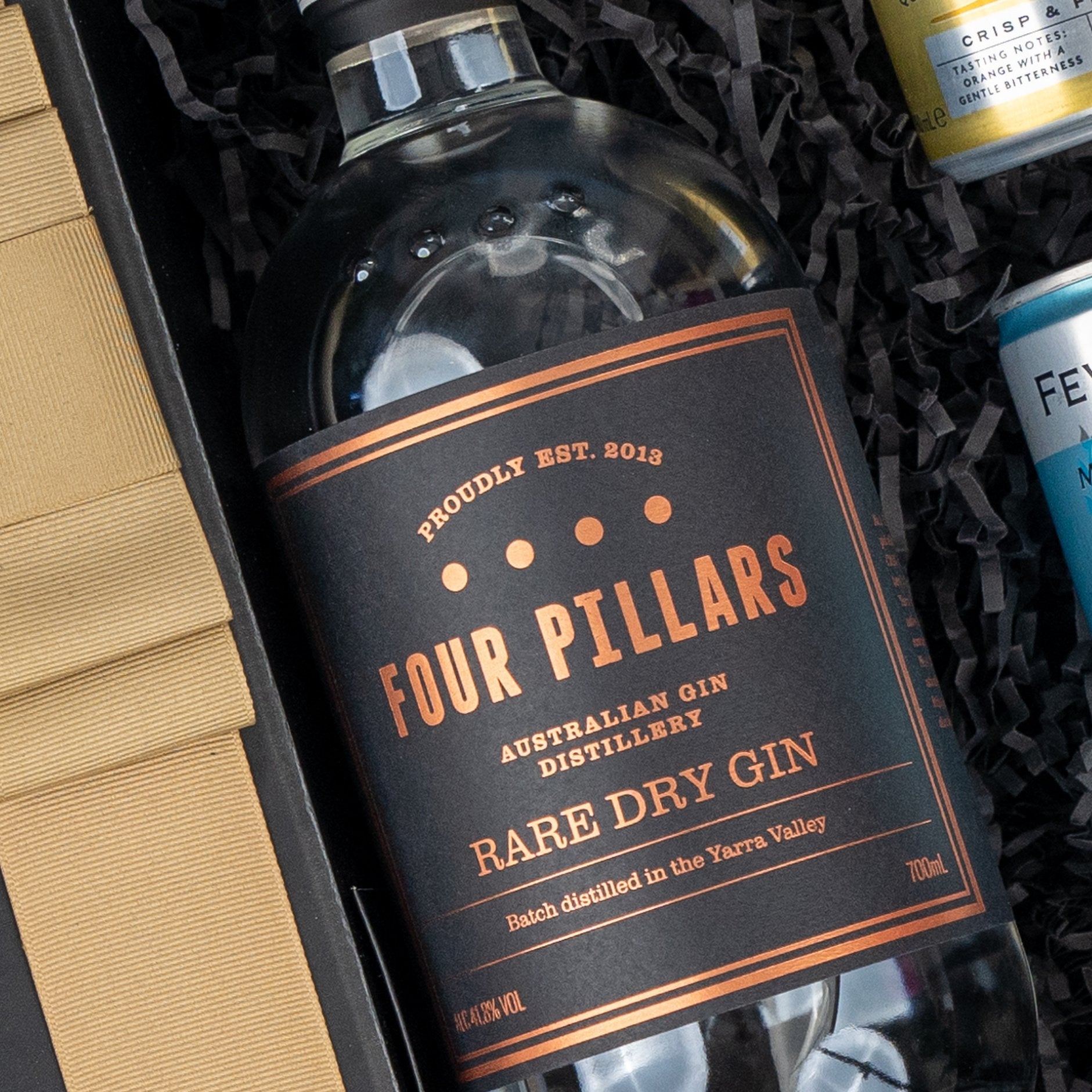 The Hamper Boutique Co’s Four Pillars Discovery Hamper features a bottle of Four Pillars Rare Dry Gin in a black and copper label, presented in a gift box with shredded black paper and assorted beverage cans.