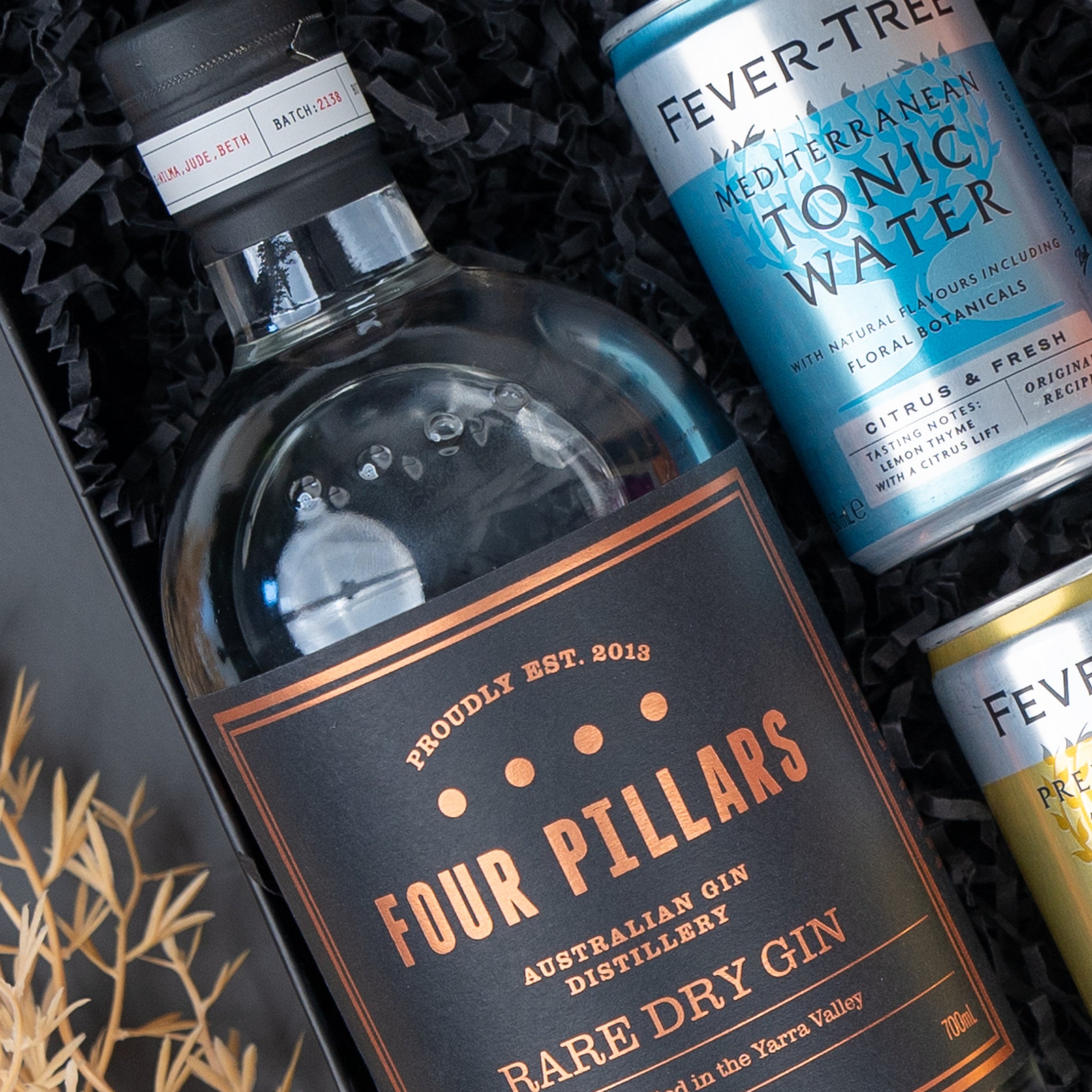 The Hamper Boutique Co's Four Pillars Signature Collection Hamper features a bottle of gin and two Fever-Tree tonics in a gift box with black shredded paper and visible decorative foliage.