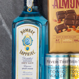 The Hamper Boutique Co's Bombay Sapphire Indulgence Hamper features a bottle of gin, Whittakers Almond Gold chocolate, and two Fever-Tree Mediterranean tonic waters displayed on a marble surface.