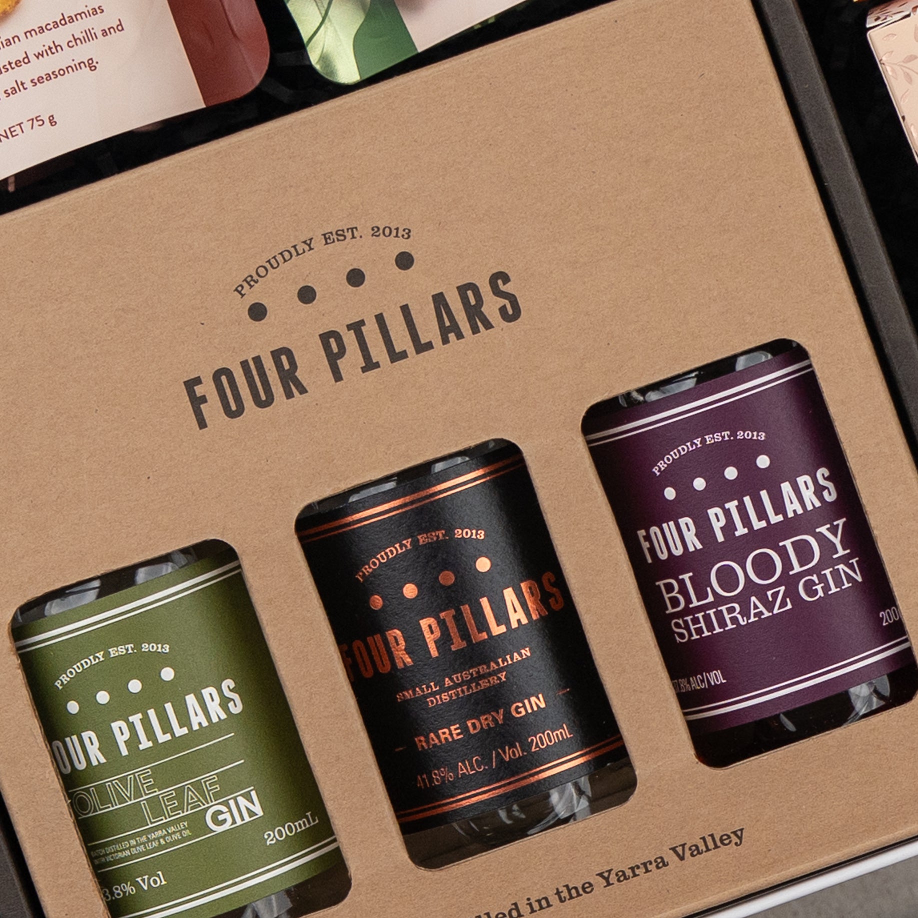 A close-up of The Hamper Boutique Co's Four Pillars Ultimate Gin Experience Hamper, featuring three 200ml bottles—Olive Leaf, Rare Dry, and Bloody Shiraz Gin—arranged in a cardboard display with visible labels.
