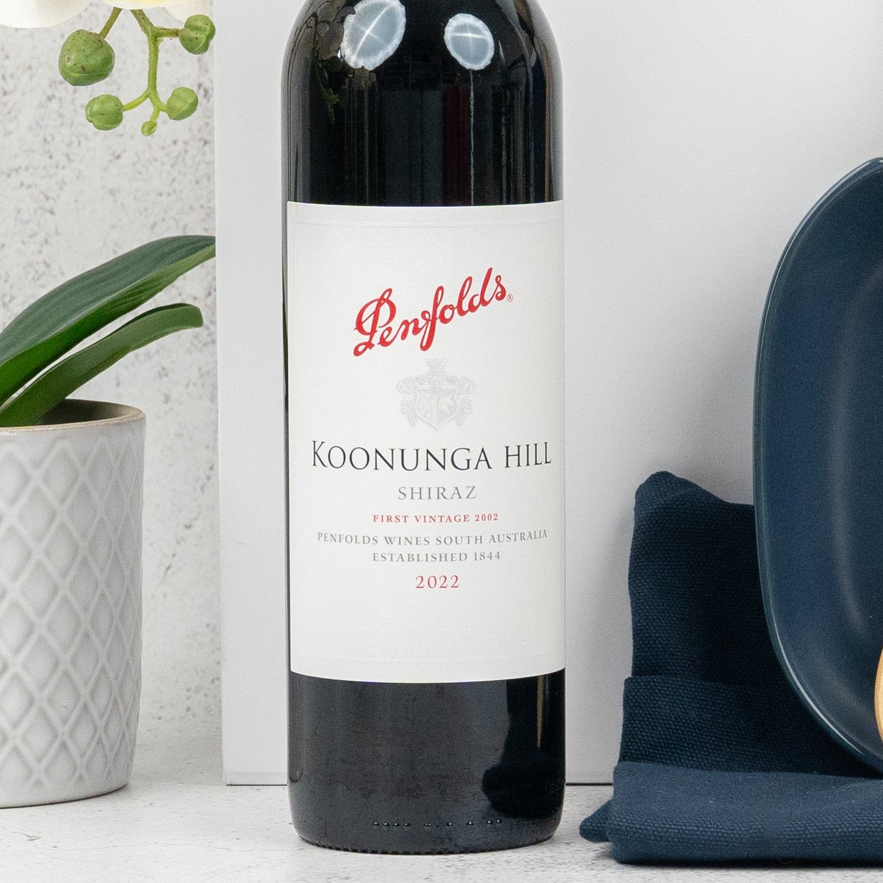 A bottle of Wine and Dine by The Hamper Boutique Co sits on a white countertop beside a green plant in a patterned white pot, a navy napkin, and a dark plate.