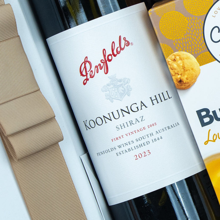 A close-up of The Hamper Boutique Co Classic Delights Gift Hamper, showing a Penfolds Koonunga Hill Shiraz 2023 wine bottle beside a gold ribbon and a yellow and white box of cookies.