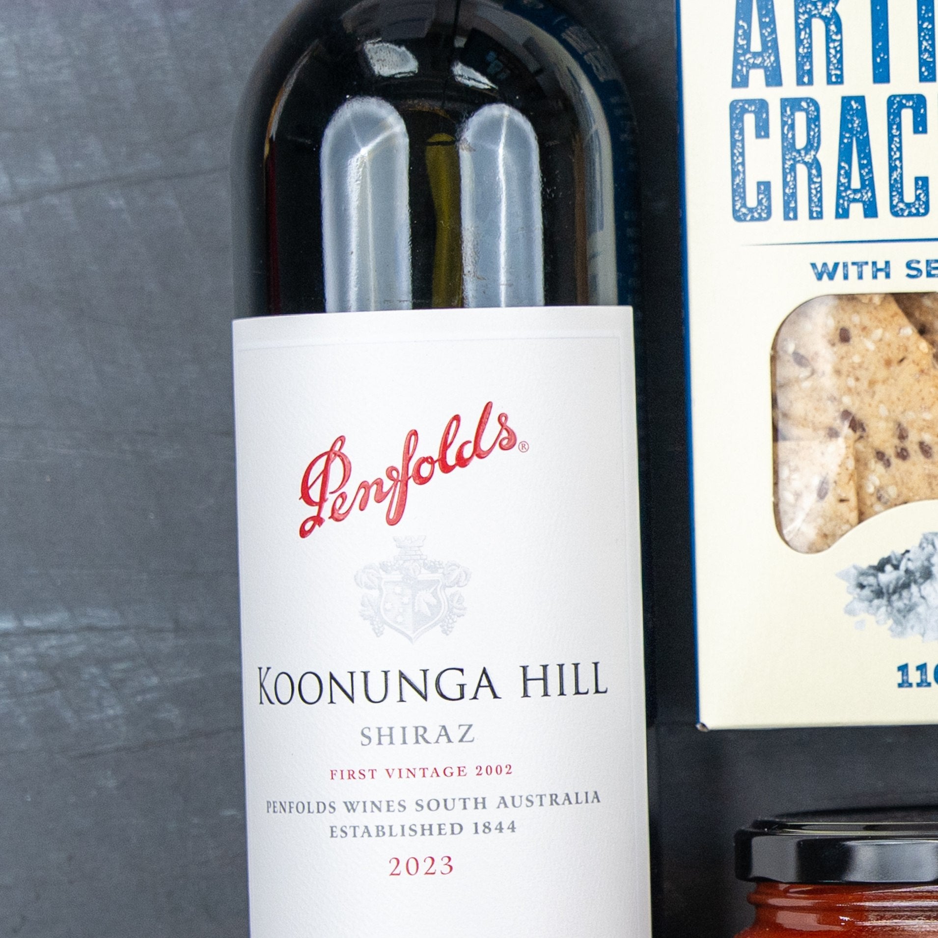 A close-up of The Hamper Boutique Co Savoury Selection Gift Hamper featuring wine, artisan crackers, and a glass jar lid arranged on a dark surface.