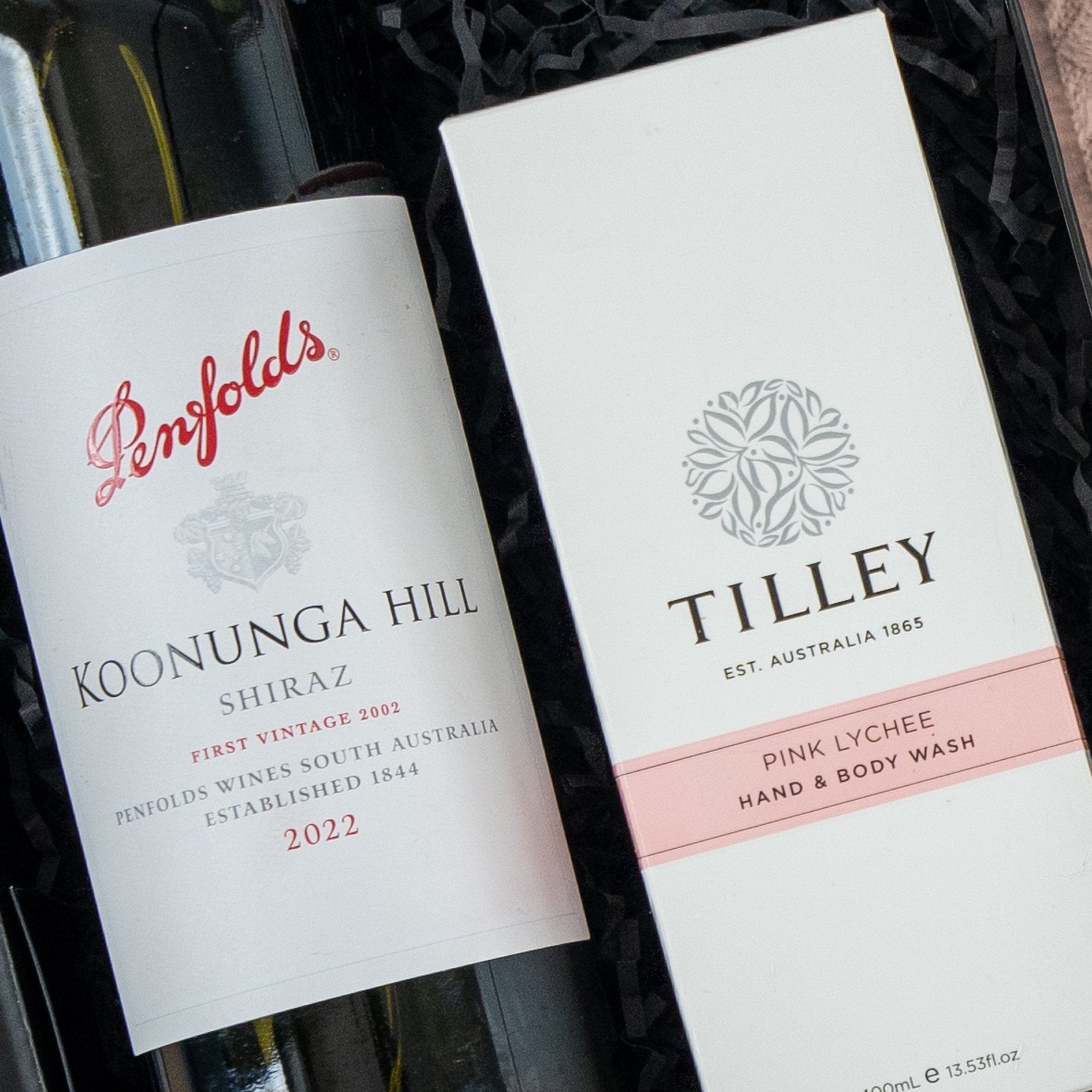 The Hamper Boutique Co's Pamper and Indulge Gift Hamper includes a bottle of Penfolds Koonunga Hill Shiraz 2022 wine and Tilley Pink Lychee Hand & Body Wash, elegantly arranged on black shredded paper.