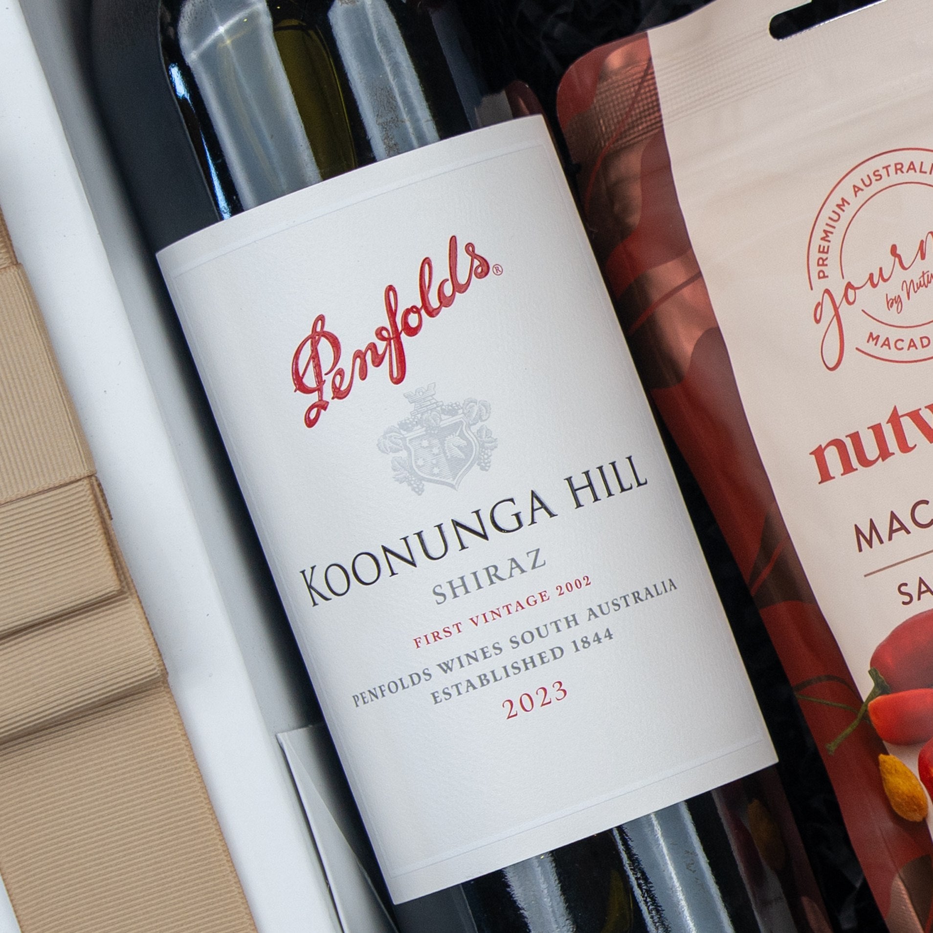 A Flavour Fiesta Gift Hamper from The Hamper Boutique Co, featuring a 2023 Penfolds Koonunga Hill Shiraz bottle beside a nut products package. The wine label has the brand name and details in red and gray text.