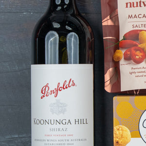 The Festive Treats Gift Hamper by The Hamper Boutique Co, featuring Penfolds Koonunga Hill Shiraz, salted macadamia nuts, and a yellow-patterned box, sits on a dark wooden surface.