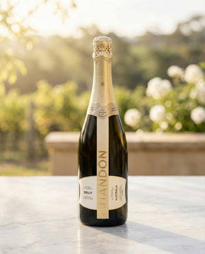 A bottle of Chandon Brut NV sparkling wine stands on a marble surface outdoors, with a blurred green garden and white flowers in the sunny background.