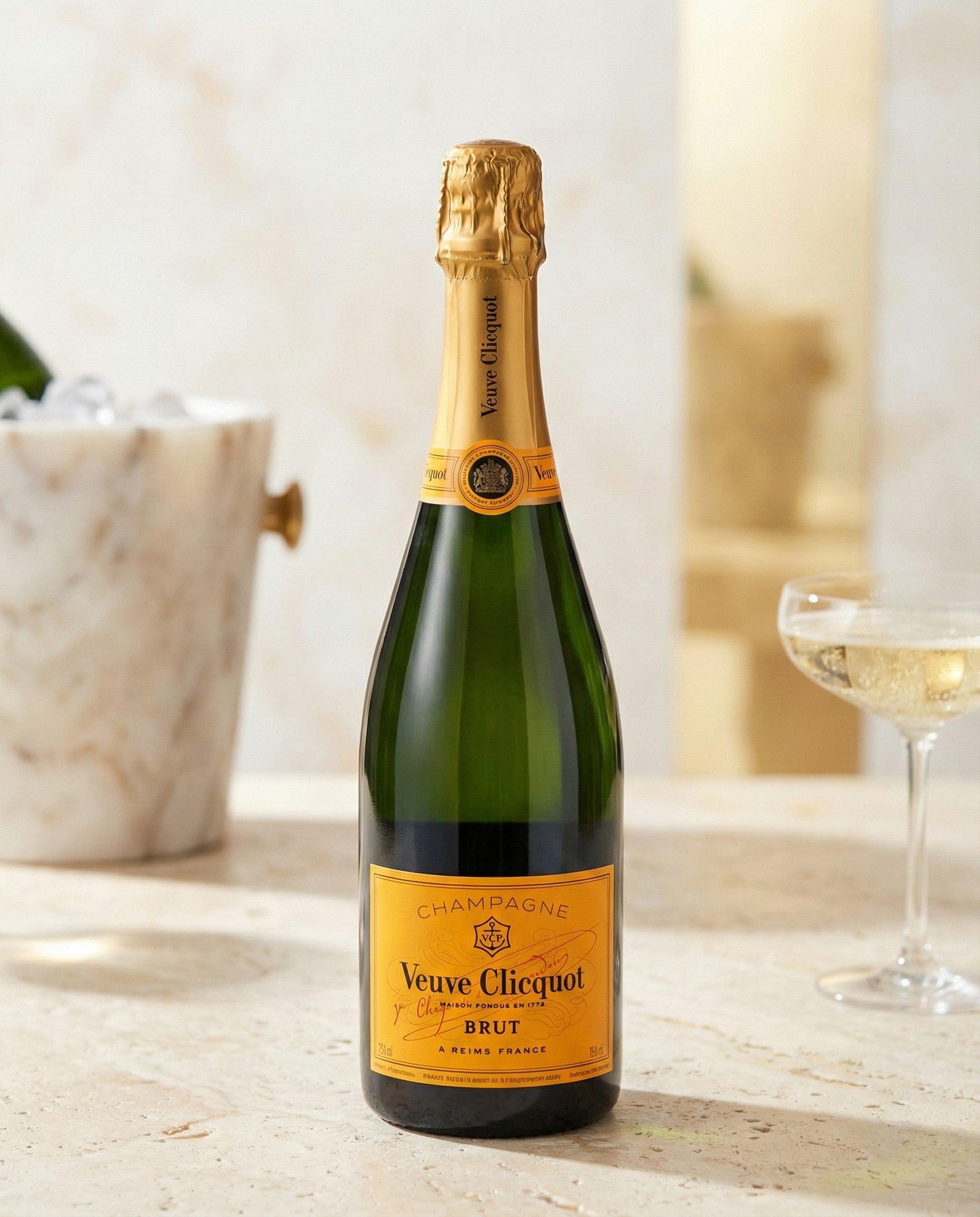 A bottle of Veuve Clicquot Brut NV sits on a light surface beside a marble ice bucket and a glass of champagne.