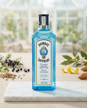 A blue bottle of Bombay Sapphire London Dry Gin stands on a marble surface, accompanied by botanicals such as juniper berries, almonds, and lemon peels, with a blurred garden in the background.