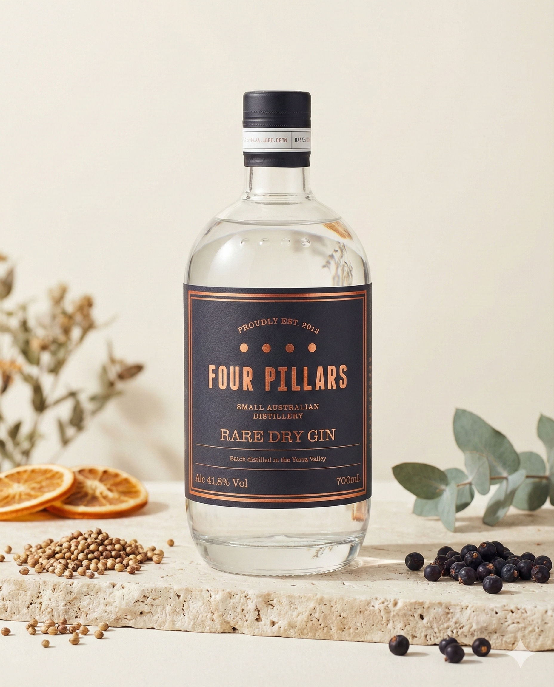 A bottle of Four Pillars Rare Dry Gin rests on a stone surface, surrounded by botanicals such as coriander seeds, juniper berries, dried citrus slices, and eucalyptus leaves with dried flowers in the background.