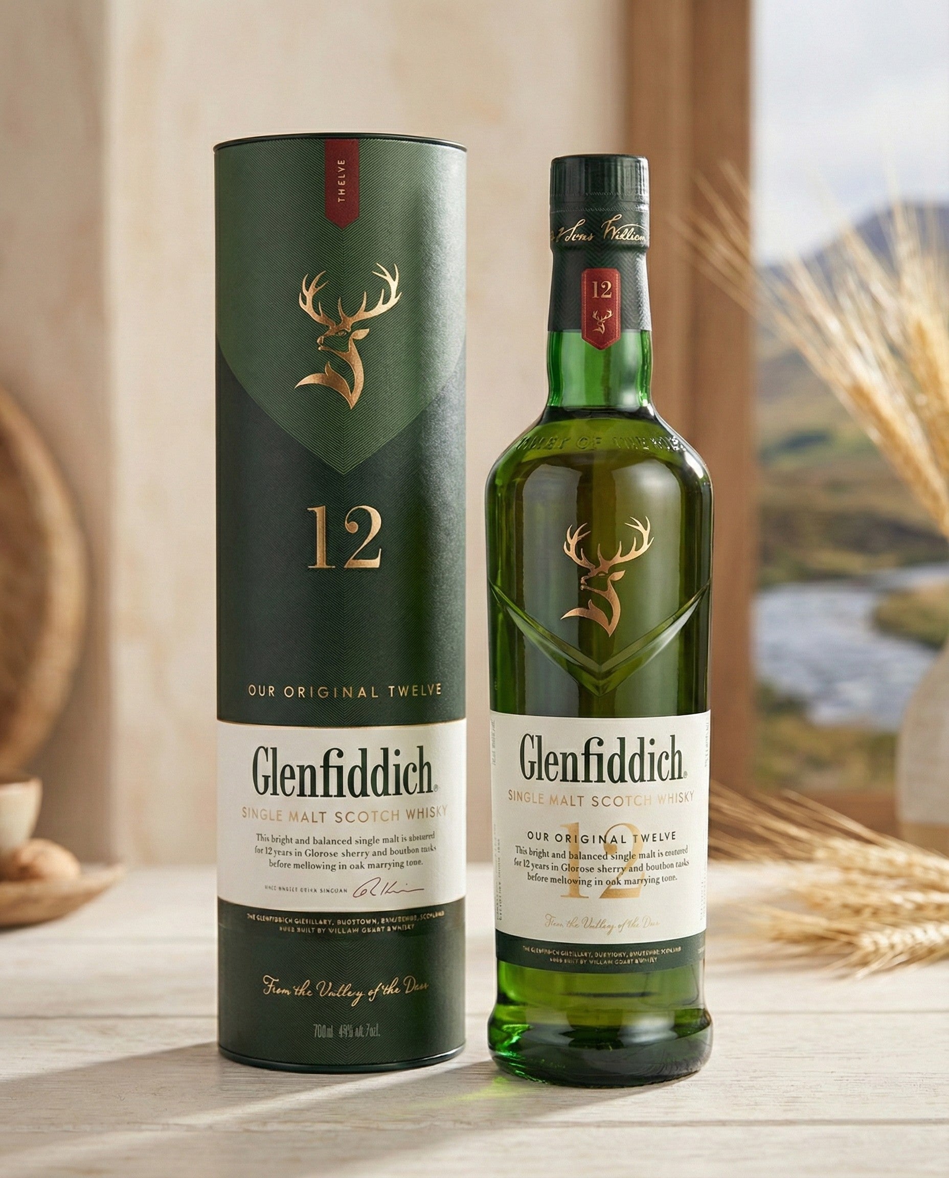 A bottle of Glenfiddich 12-Year-Old Whisky is displayed beside its green cylindrical box on a light wood table, with wheat stalks and a softly blurred landscape in the background.