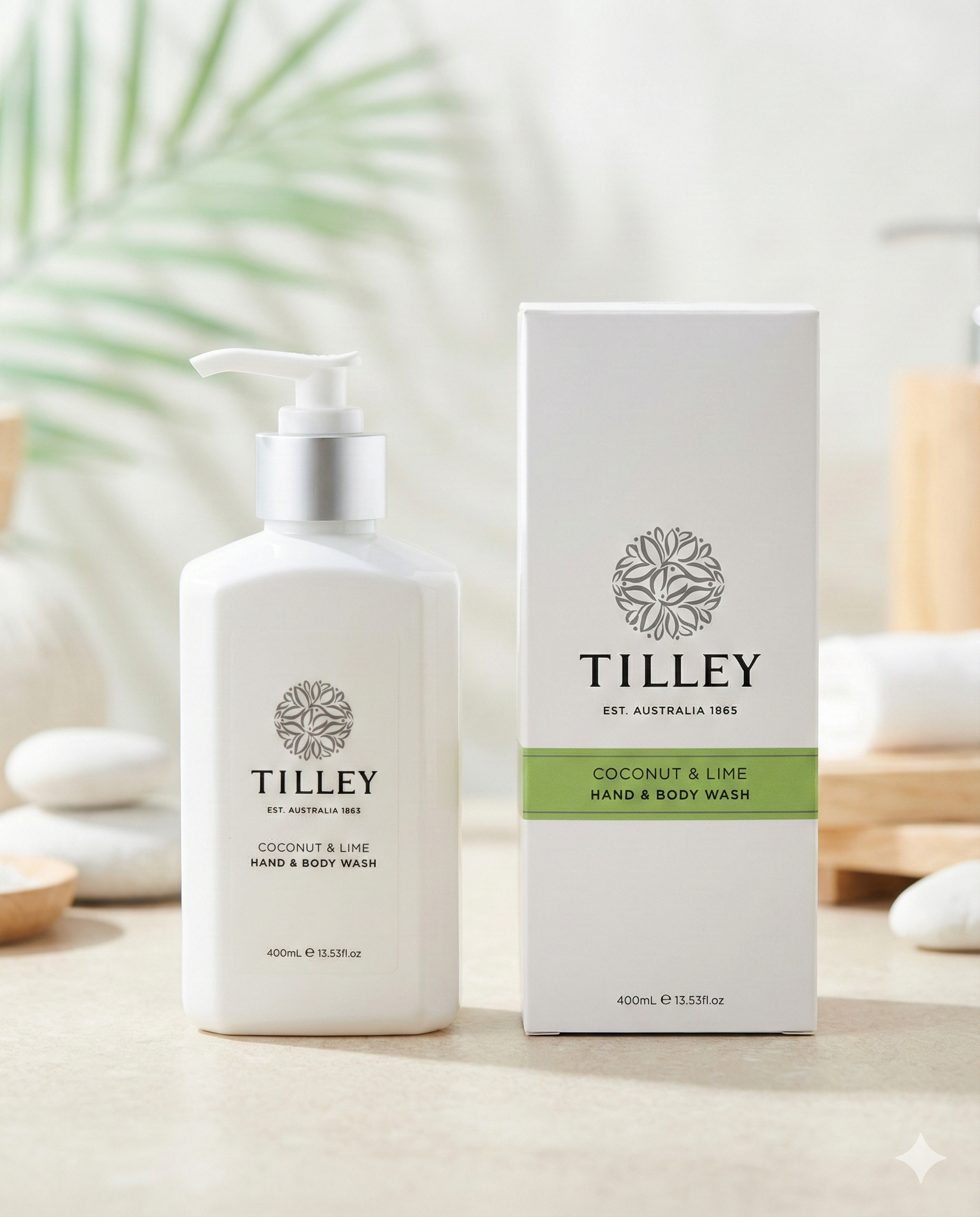 A white pump bottle labeled Tilley Coconut and Lime Body Wash stands beside its matching box on a light surface, with smooth stones and soft-focus greenery in the background.