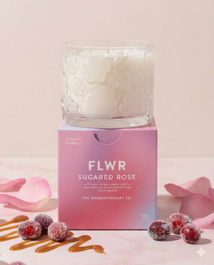 The Aromatherapy Company's FLWR Sugared Rose Candle rests on its pink box, surrounded by caramel drizzles, sugared cranberries, and pink rose petals atop a soft pink surface.