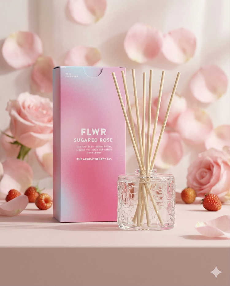 A pink and blue box of The Aromatherapy Company’s Flwr Sugared Rose Diffuser stands next to a glass jar with reed sticks, surrounded by pink roses, rose petals, and small strawberries on a pale surface.