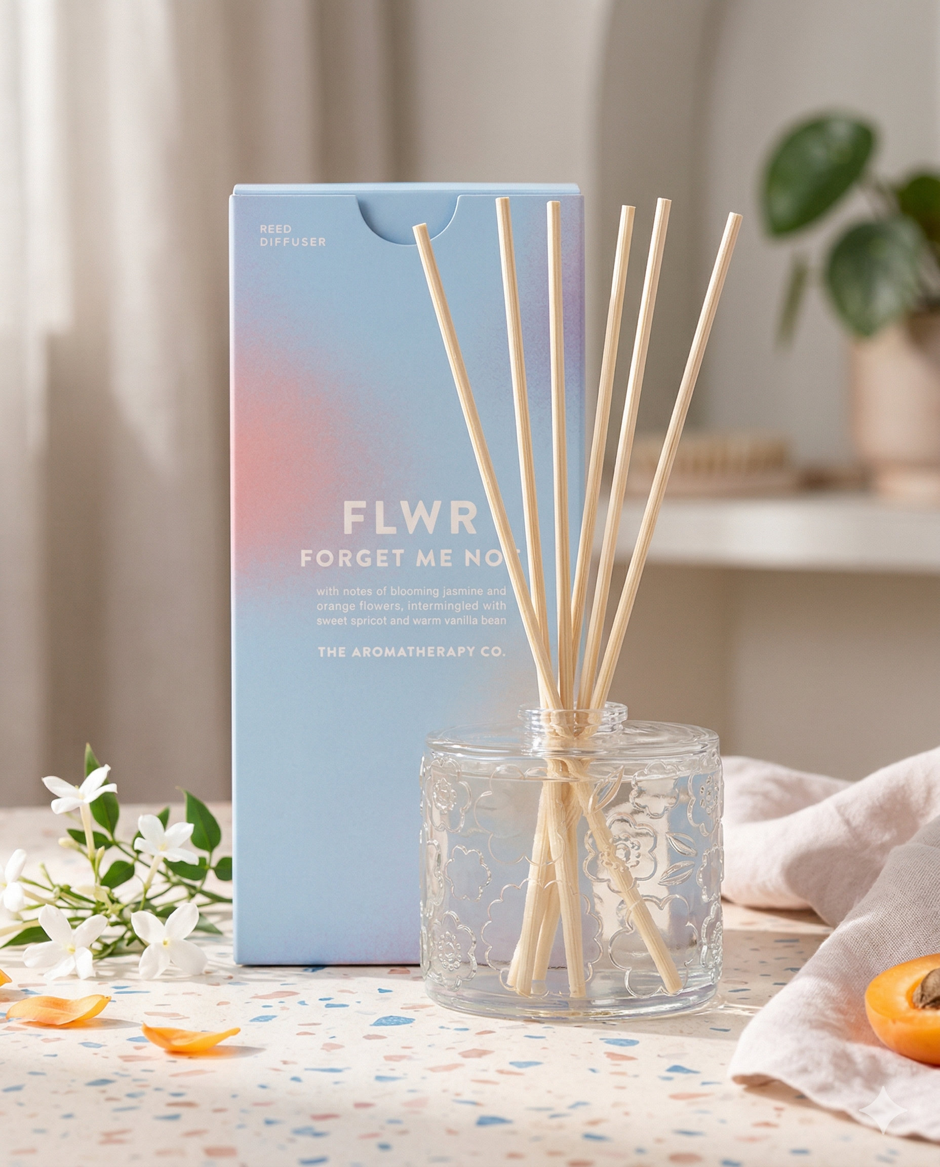 A glass Flwr Forget Me Not Diffuser by The Aromatherapy Company with light wooden sticks stands before its pastel box, surrounded by white flowers, petals, and an orange slice on a softly lit table.