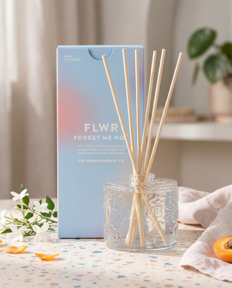 A glass Flwr Forget Me Not Diffuser by The Aromatherapy Company with light wooden sticks stands before its pastel box, surrounded by white flowers, petals, and an orange slice on a softly lit table.