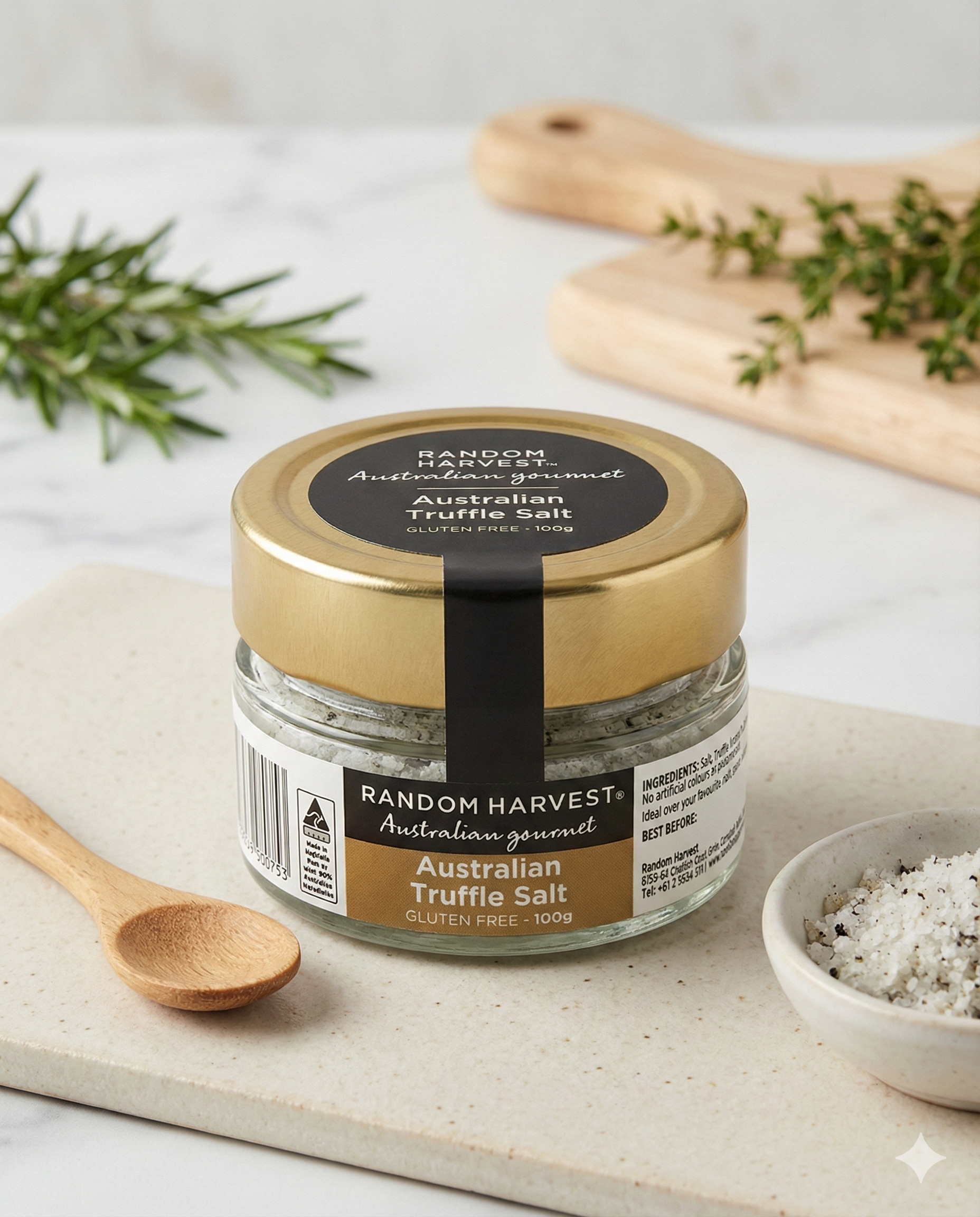 A jar of Random Harvest Australian Truffle Salt rests on a white surface with a small wooden spoon, a pinch bowl of salt, fresh herbs, and a wooden board topped with chopped herbs in the background.