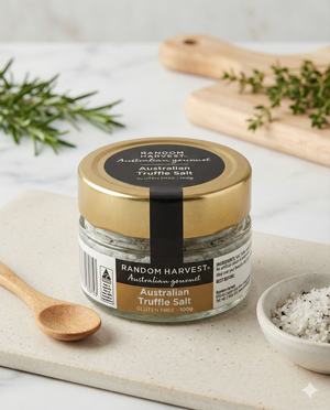 A jar of Random Harvest Australian Truffle Salt rests on a white surface with a small wooden spoon, a pinch bowl of salt, fresh herbs, and a wooden board topped with chopped herbs in the background.