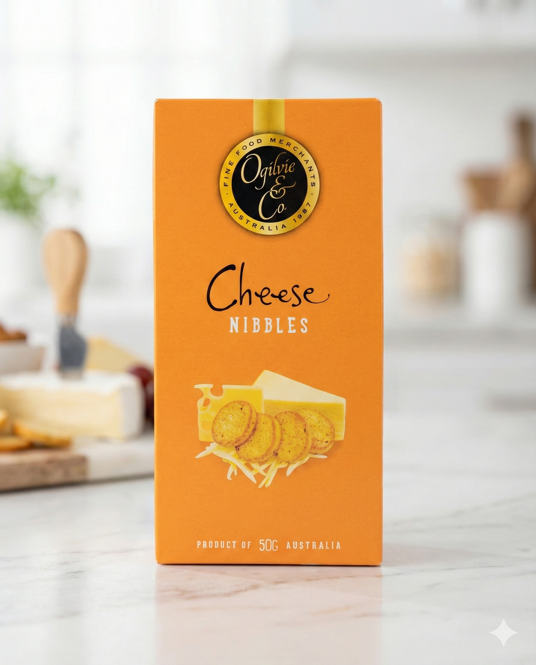 An orange box of Ogilvie and Co Cheese Nibbles rests on a white kitchen counter, with cheese and other kitchen items blurred in the background. The packaging features images of crackers and shredded cheese.