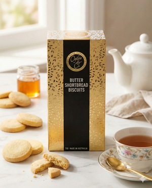 A gold and black box of Ogilvie and Co Golden Butter Shortbread sits on a table with a teacup, teapot, honey jar, and several shortbread biscuits beside it.