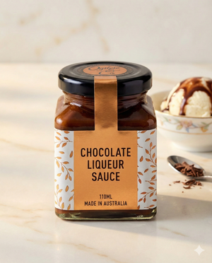 A jar of Ogilvie and Co Chocolate Liqueur Sauce (110ml, Made in Australia) sits on marble, with a bowl of vanilla ice cream topped with the sauce and chocolate shavings on a spoon in the background.