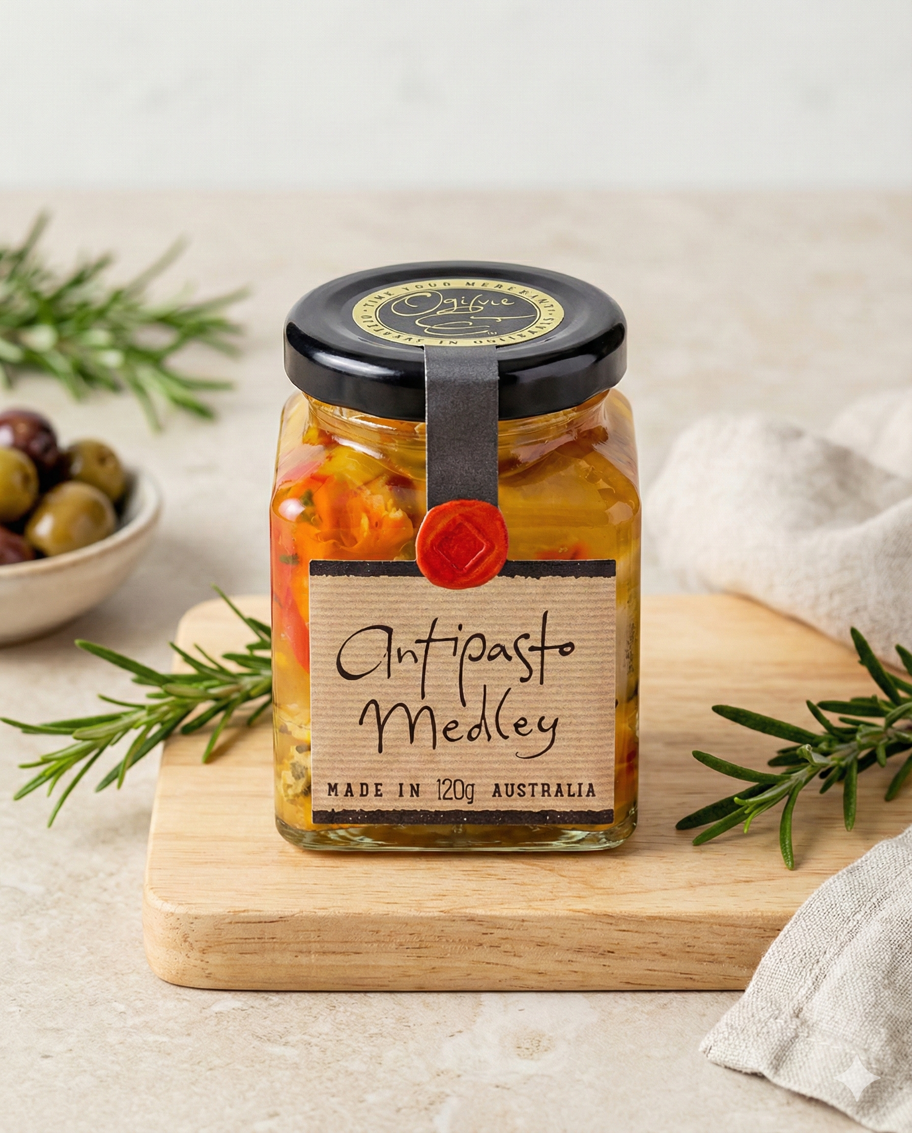 A jar of Ogilvie and Co Antipasto Medley sits on a wooden board with rosemary, a bowl of olives, and a beige napkin. The black-lidded jar holds colorful preserved vegetables.