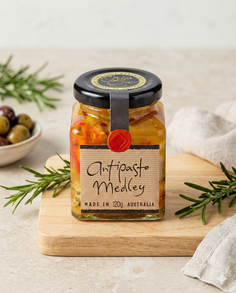 A jar of Ogilvie and Co Antipasto Medley sits on a wooden board with rosemary, a bowl of olives, and a beige napkin. The black-lidded jar holds colorful preserved vegetables.