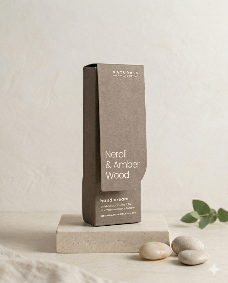 A box of The Aromatherapy Company Naturals Hand Cream Neroli and Amber Wood rests on a stone slab, with smooth pebbles and a green plant in the background against a light, neutral setting.