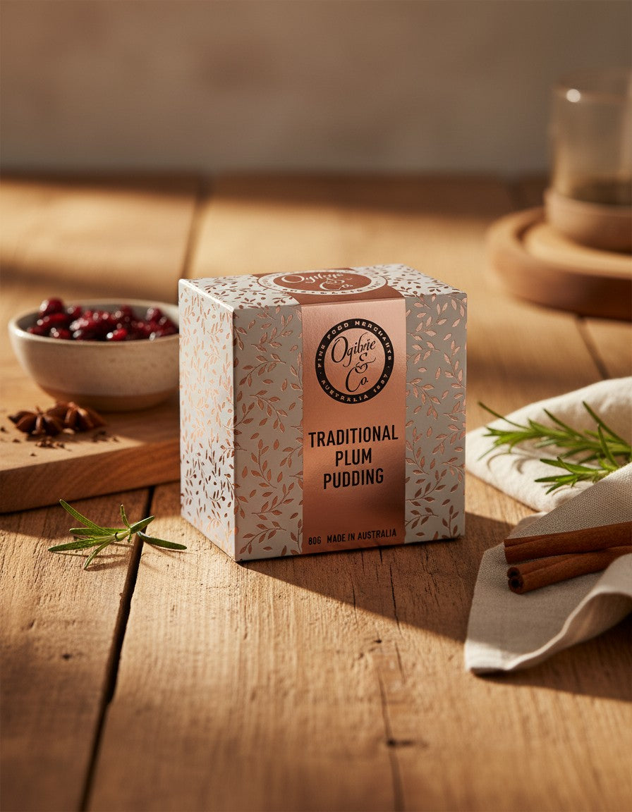A box of Ogilvie and Co Plum Pudding rests on a wooden table, accompanied by a bowl of red berries, cinnamon stick, star anise, and sprigs of rosemary, all bathed in warm sunlight.