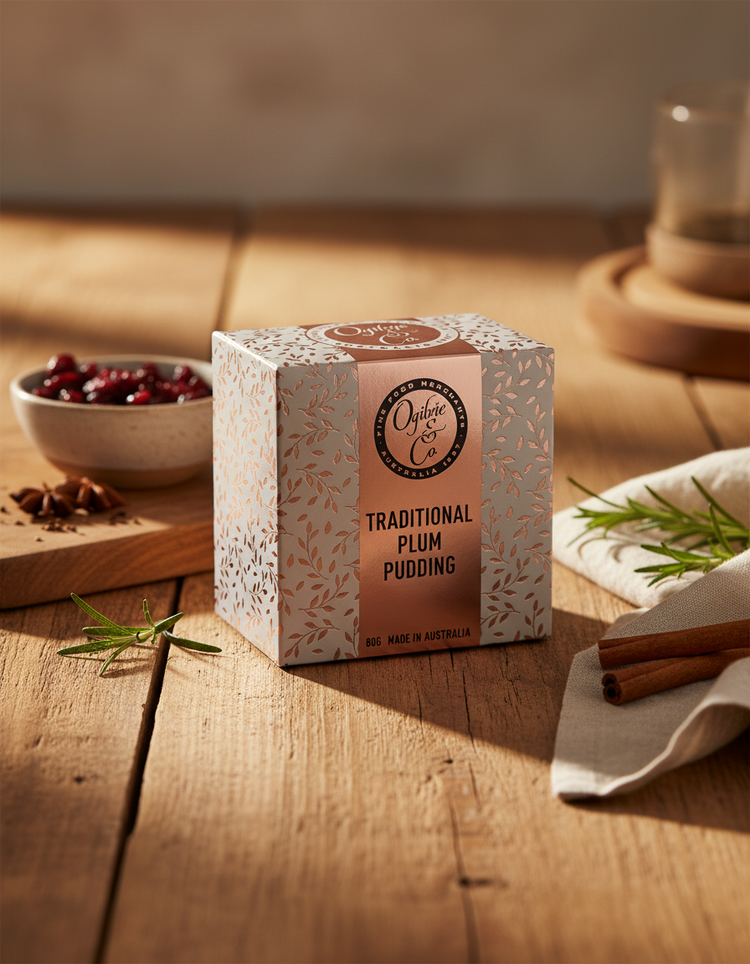 An Ogilvie and Co Plum Pudding rests on a wooden table, accompanied by cinnamon sticks, rosemary, a napkin, and cranberries. Warm sunlight adds to the cozy ambiance.