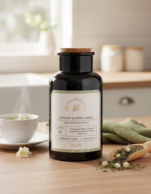 A black tea canister from The Tea Collective, labeled Jasmine Flower Green, sits on a wooden table alongside a steaming teacup, loose tea leaves, fresh white jasmine flowers, and a green napkin. Sunlight streams through a window in the background.