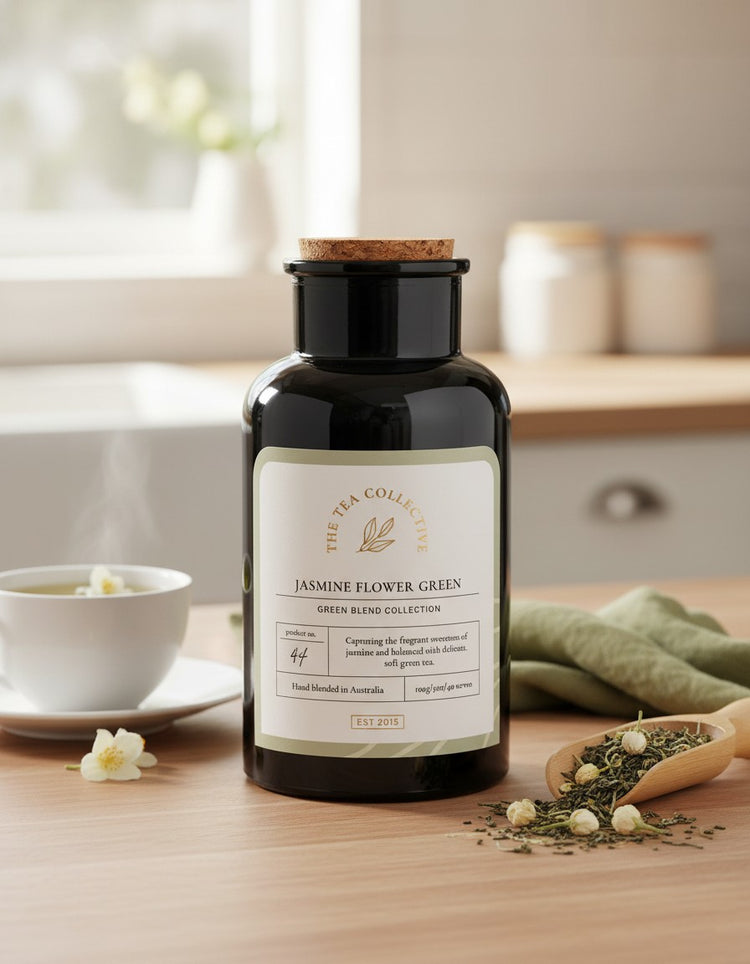 A black tea canister from The Tea Collective, labeled Jasmine Flower Green, sits on a wooden table alongside a steaming teacup, loose tea leaves, fresh white jasmine flowers, and a green napkin. Sunlight streams through a window in the background.