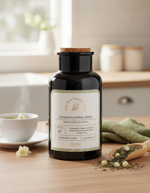 A dark glass jar of The Tea Collective's Jasmine Flower Green sits on a wooden table beside a steaming teacup, loose tea leaves, and jasmine flowers, creating a calm and inviting tea scene.
