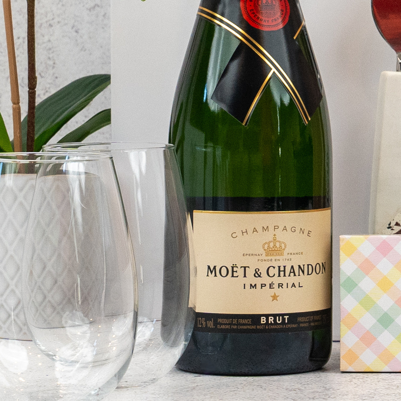 A bottle from The Hamper Boutique Co's Champagne and Homeware Collection sits on a countertop beside two empty wine glasses, a potted plant, and a pastel checkered gift box.
