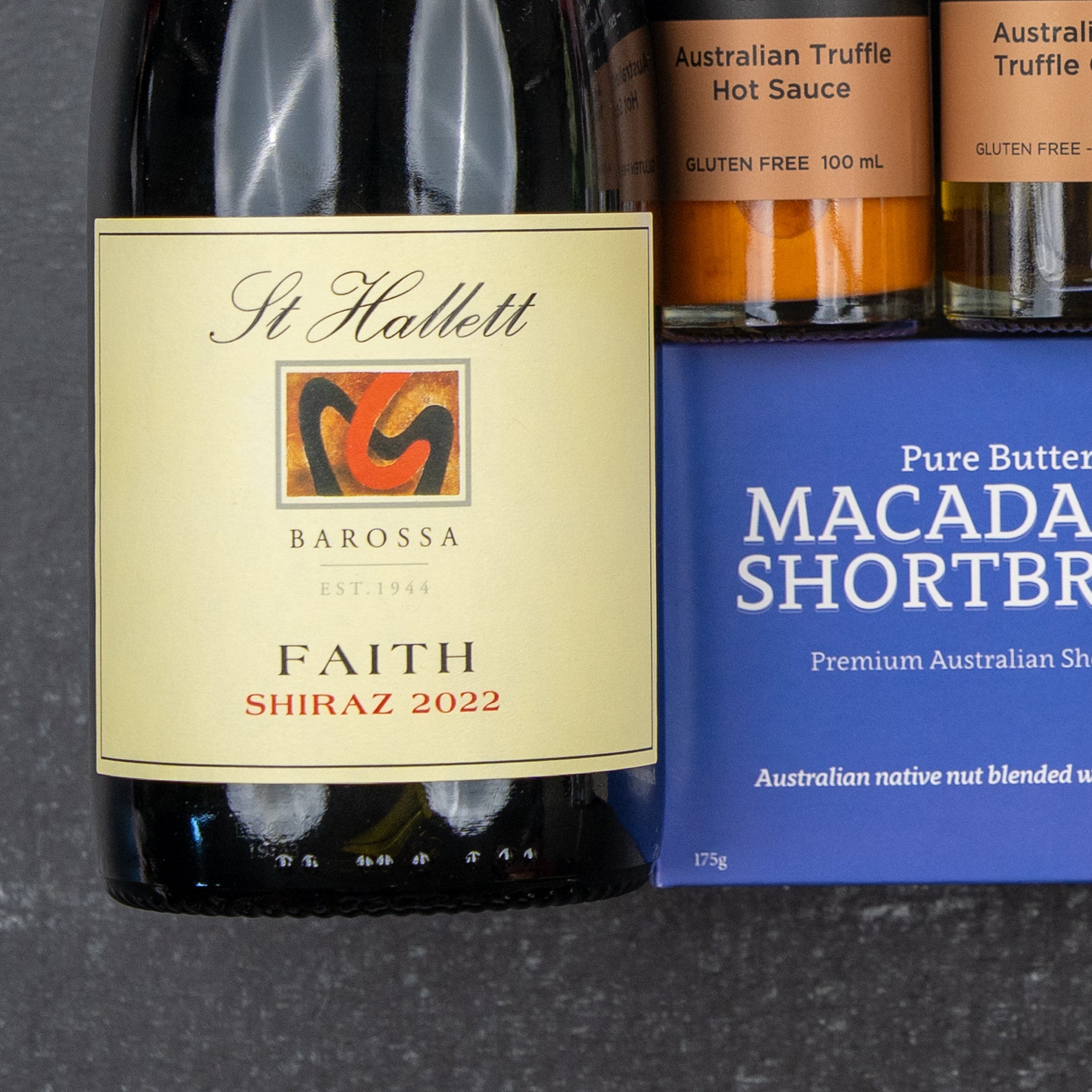 The Hamper Boutique Co Luxury Indulgence Gift Hamper featuring wine, Australian truffle hot sauce, and macadamia shortbread is elegantly arranged on a dark surface.