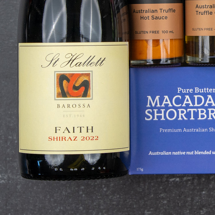 The Hamper Boutique Co Luxury Indulgence Gift Hamper featuring wine, Australian truffle hot sauce, and macadamia shortbread is elegantly arranged on a dark surface.