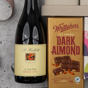 An Indulgent Evening Escape hamper from The Hamper Boutique Co sits on a grey surface, featuring wine and chocolate with other products partially visible nearby.