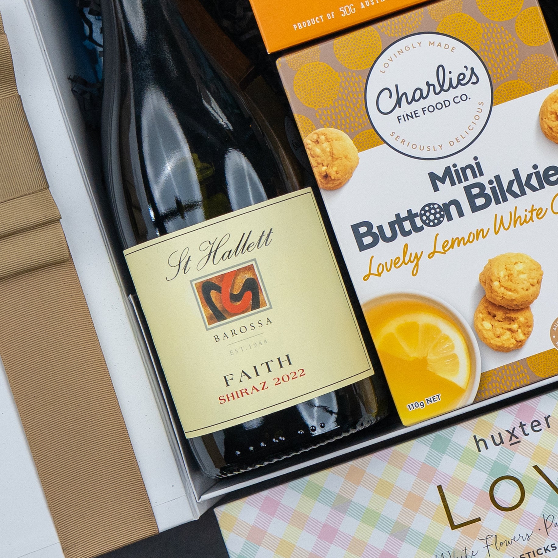 The Hamper Boutique Co's Red Wine Culinary Experience Hamper includes a bottle of St Hallett Faith Shiraz 2022, Charlie’s Mini Button Bikkies Lovely Lemon White Choc biscuits, and a floral-themed Huxter soap bar.